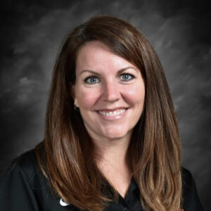 Smiling woman with long brown hair, wearing a black shirt, against a dark gray background.