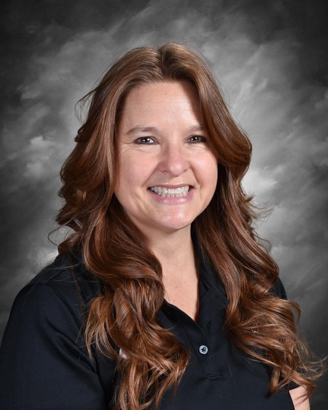 Smiling woman with long, wavy brown hair wearing a black shirt, set against a gray backdrop.