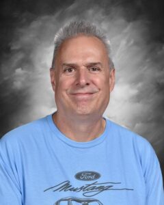 A smiling man with short gray hair, wearing a light blue Ford Mustang t-shirt, poses against a dark background.