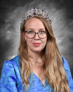 A woman with long hair wears a jeweled crown and a blue blouse, posing against a gray background.