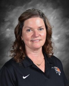 Portrait of a woman with curly hair, wearing a black polo shirt with a tiger logo, smiling against a gray background.