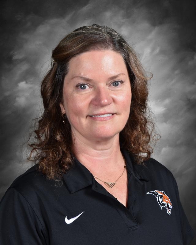 Portrait of a woman with curly hair, wearing a black polo shirt with a tiger logo, smiling against a gray background.