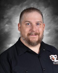 Smiling man with a beard, wearing a black shirt, against a gray background. He appears friendly and approachable.