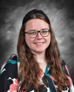 Young woman with long, wavy hair and glasses, wearing a floral blouse, smiling against a gray backdrop.