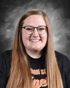 A smiling young woman with long hair wearing glasses and a black t-shirt with "High School Tigers" printed on it.