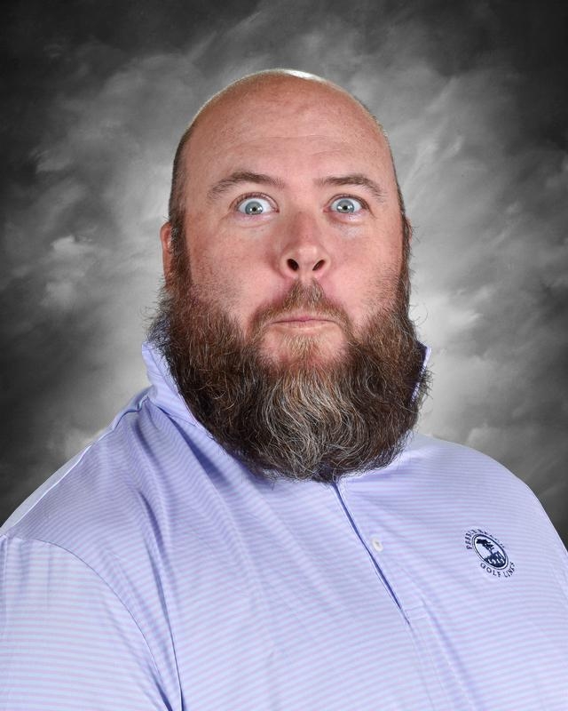 A man with a large beard makes a surprised expression, wearing a light purple collared shirt against a gray backdrop.
