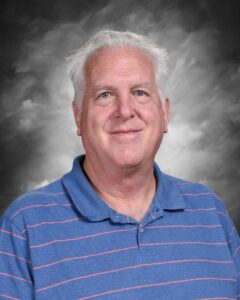 Smiling man with gray hair wearing a blue striped polo shirt, against a soft gray background.