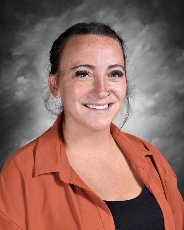 Smiling woman with brown hair and a nose ring, wearing an orange shirt, against a gray background.