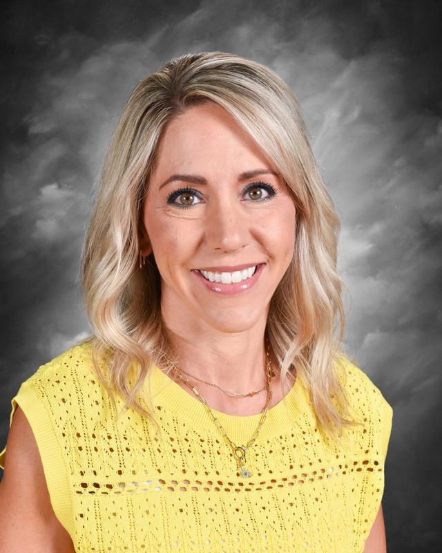 A smiling woman with blonde hair wearing a yellow sweater, set against a gray background.