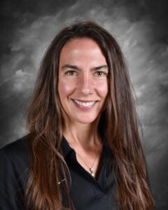 A smiling woman with long brown hair wears a black shirt, set against a gray background.