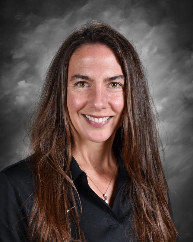 A smiling woman with long brown hair wears a black shirt, set against a gray background.