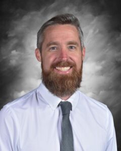Smiling man with a beard wearing a white shirt and gray tie, set against a cloudy gray background.