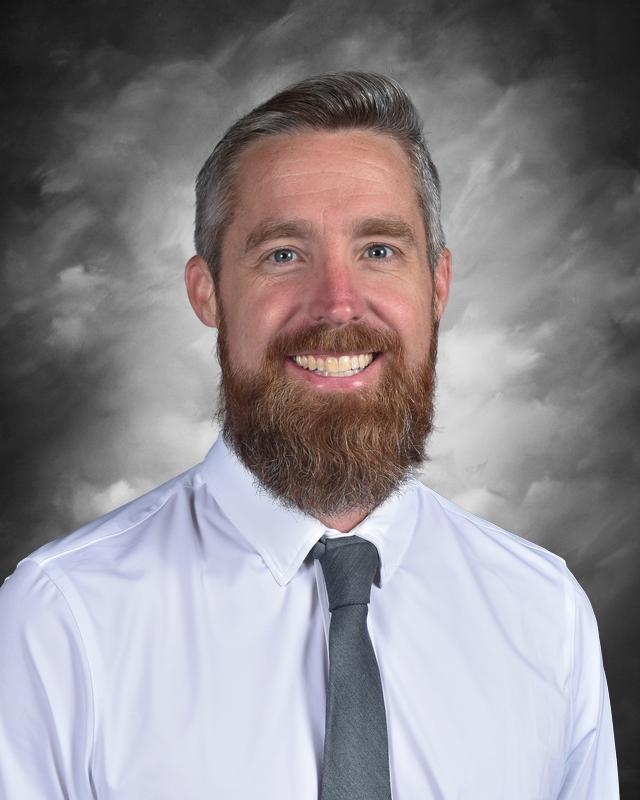Smiling man with a beard wearing a white shirt and gray tie, set against a cloudy gray background.