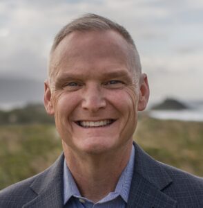 Smiling man in a suit against a scenic backdrop with ocean and hills, conveying a friendly and approachable vibe.