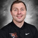 A man with short brown hair and blue eyes smiles while wearing a black polo shirt with an orange logo.