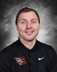 A man with short brown hair and blue eyes smiles while wearing a black polo shirt with an orange logo.