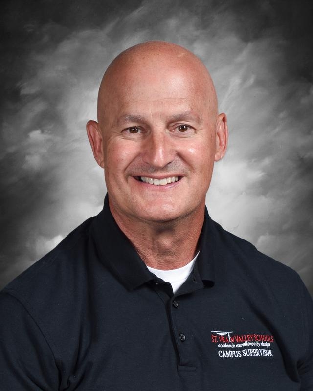 A smiling bald man in a black polo shirt with a school logo, against a gray background.