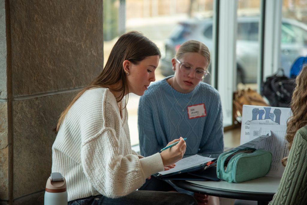 Two women are seated together at a table, focused on a notebook and discussing. One wears a white sweater, the other a blue one.