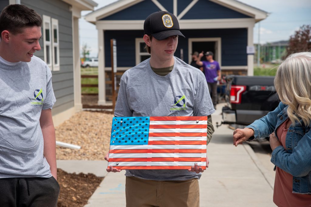 Two young men stand outside a house, one holding a metal American flag, while a woman gestures to him.