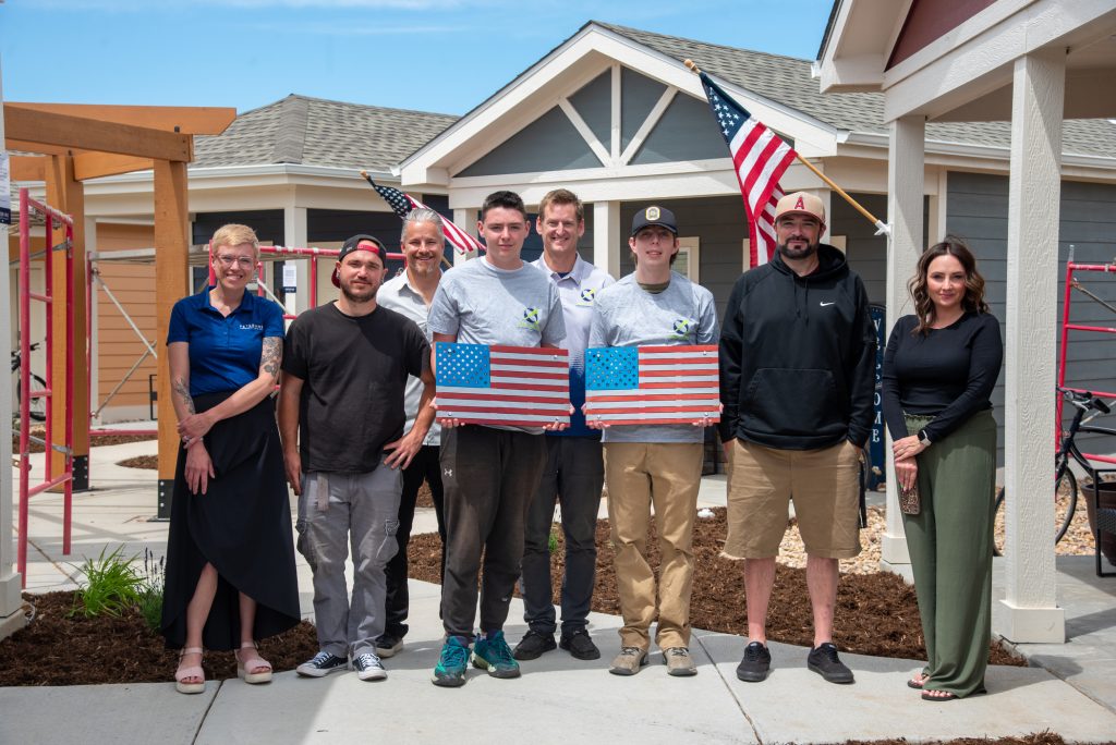 A group of eight people stands in front of a house, some holding U.S. flags, with construction scaffolding nearby.