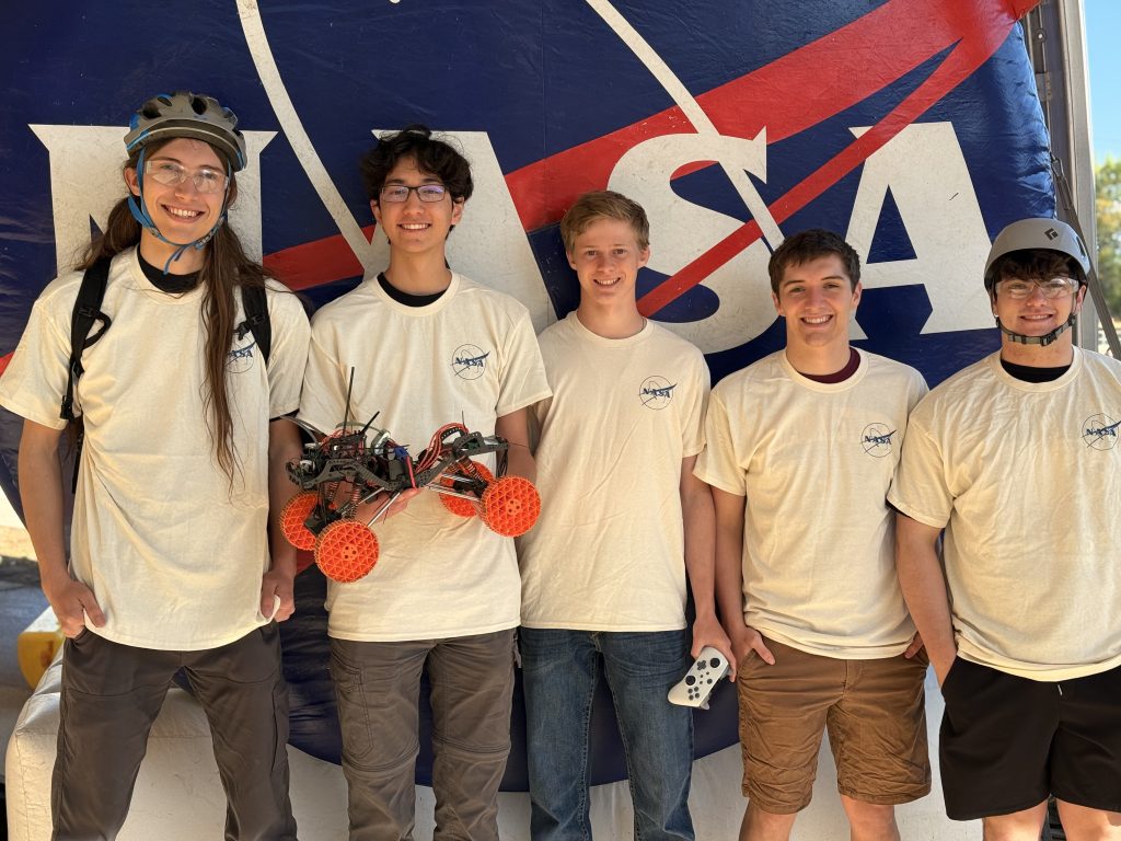 Five students in NASA t-shirts stand together, holding a robot on wheels, with a NASA banner in the background.