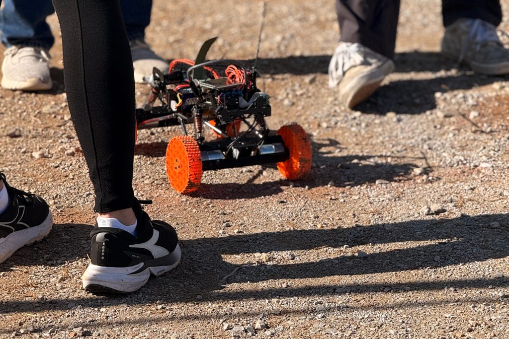 A small, remote-controlled car with bright orange wheels is on a gravel surface, near people's feet.