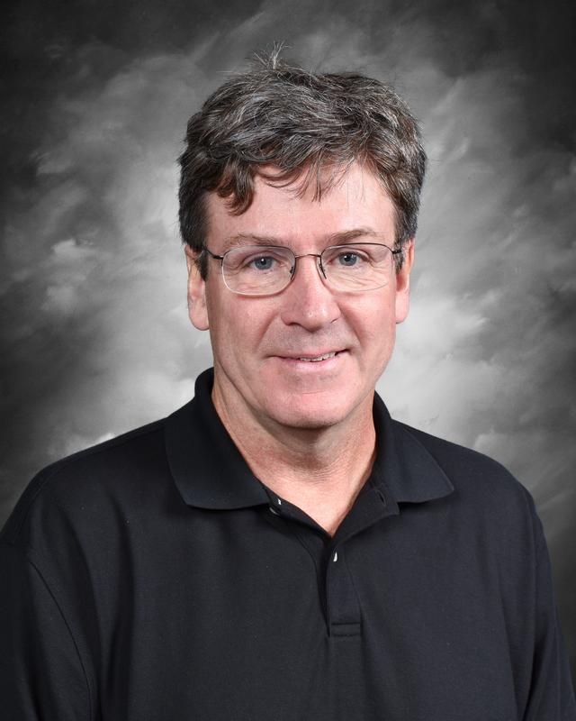 A middle-aged man with glasses and short hair smiles, wearing a black polo shirt against a gray backdrop.
