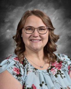 A woman with wavy hair smiles, wearing glasses and a floral blouse, against a gray background.