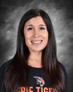 A woman with long dark hair smiles, wearing a black shirt with an orange tiger logo and text that reads "Erie Tigers."