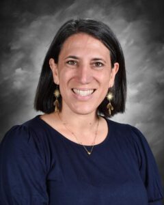 Smiling woman with short black hair, wearing a navy top and gold earrings, against a gray background.