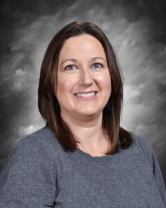 A woman with shoulder-length brown hair smiles, wearing a gray sweater, against a gray backdrop.