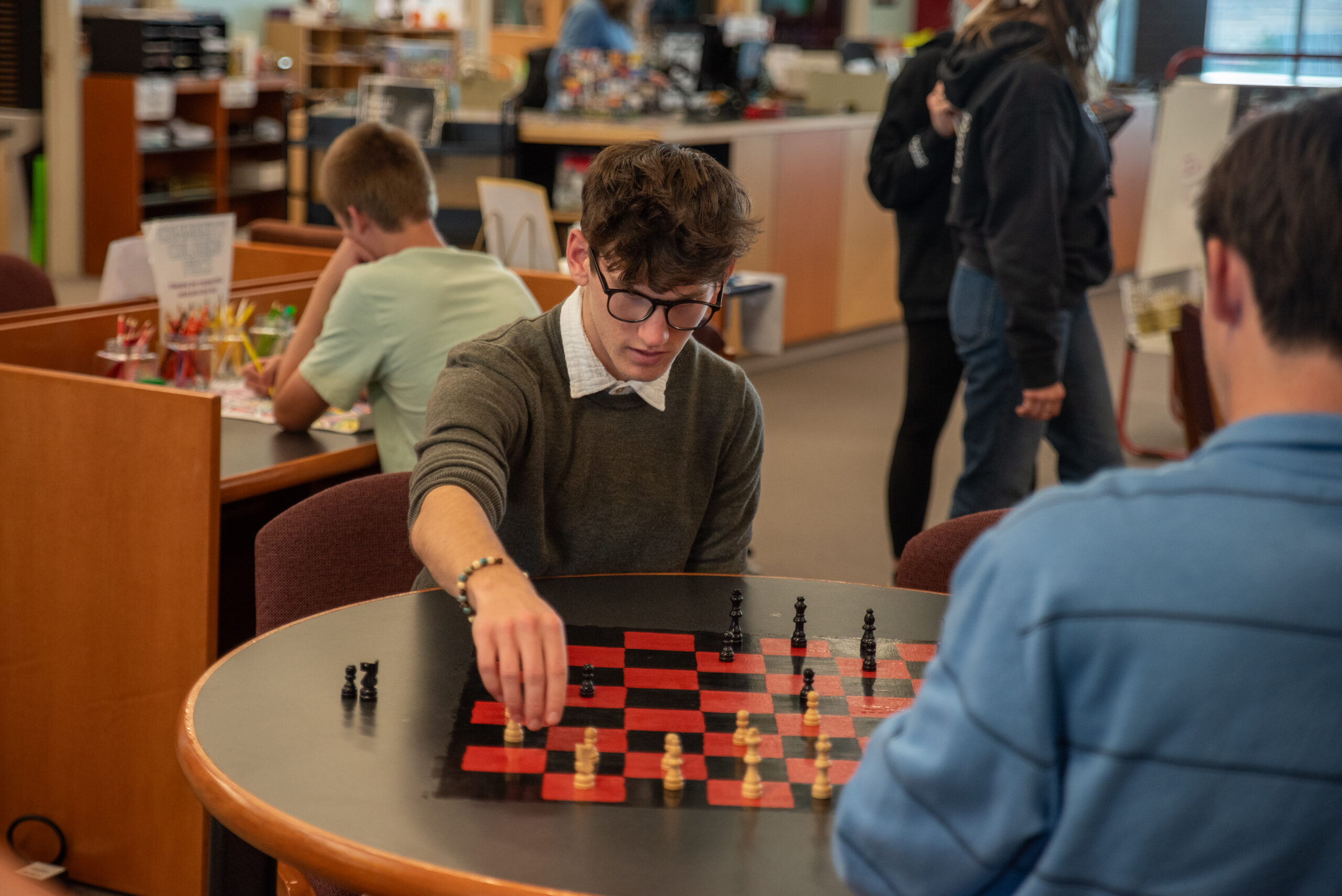 Two young men play chess at a table in a library, with other patrons engaged in various activities nearby.